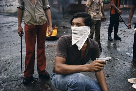 Susan Meiselas - A man in a bandana holds a pistol while other armed men are behind him. There is a tire on fire in the background. 